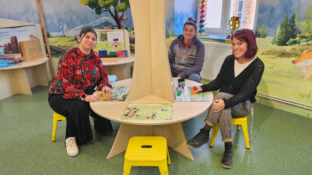 Trois femmes assises autour d'une table dans un espace éducatif coloré, avec des livres et des jeux autour d'elles.