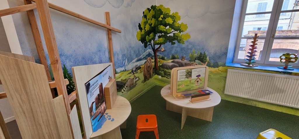 Espace éducatif avec des illustrations murales de la nature, des meubles en bois, et un coin lecture pour enfants.