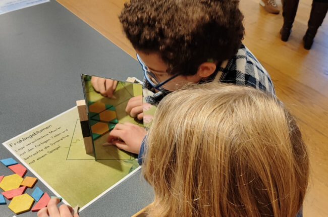 Deux enfants jouant avec des formes colorées sur une table, utilisant un miroir pour explorer les angles et les motifs.