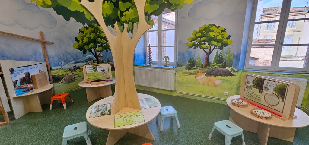 Salle d'activités pour enfants avec des tables en bois, des tabourets colorés et un décor mural représentant des arbres et des animaux. Le mobilier encourage l'interaction et l'apprentissage.